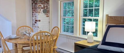 A room with a wooden ceiling, a bed with a dark cover, a dining table with chairs, and a window with a view of trees.