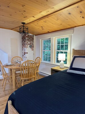 Een kamer met een houten plafond, een bed met een donkere dekbedovertrek, een eettafel met stoelen, en een raam met uitzicht op bomen.