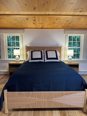 Een slaapkamer met een houten plafond, een groot bed met een donkere sprei, twee nachtkastjes met lampen, en ramen met uitzicht op bomen.