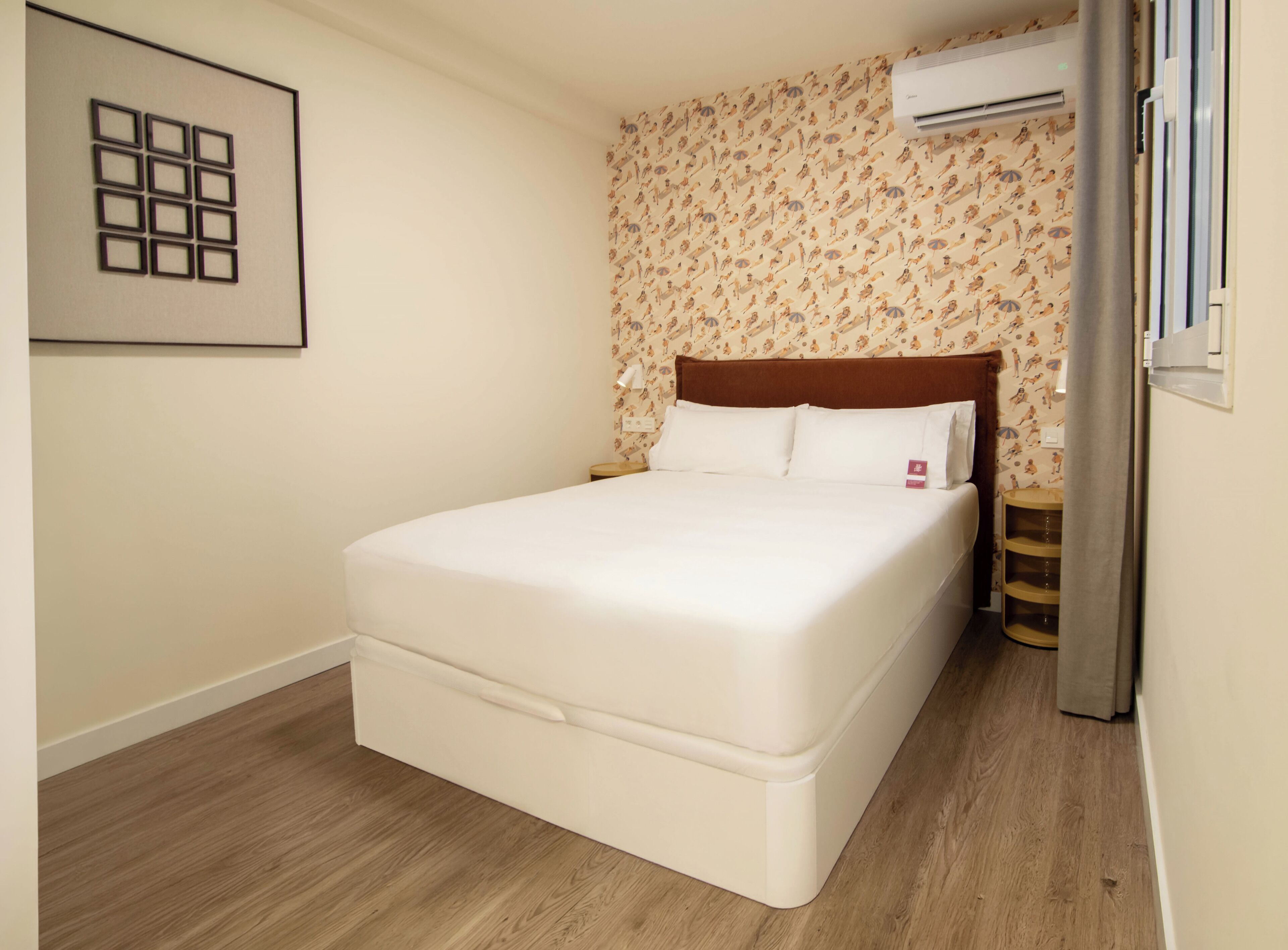 A neatly made bed in a room with a patterned wallpaper, a wooden floor, and a framed picture on the wall.