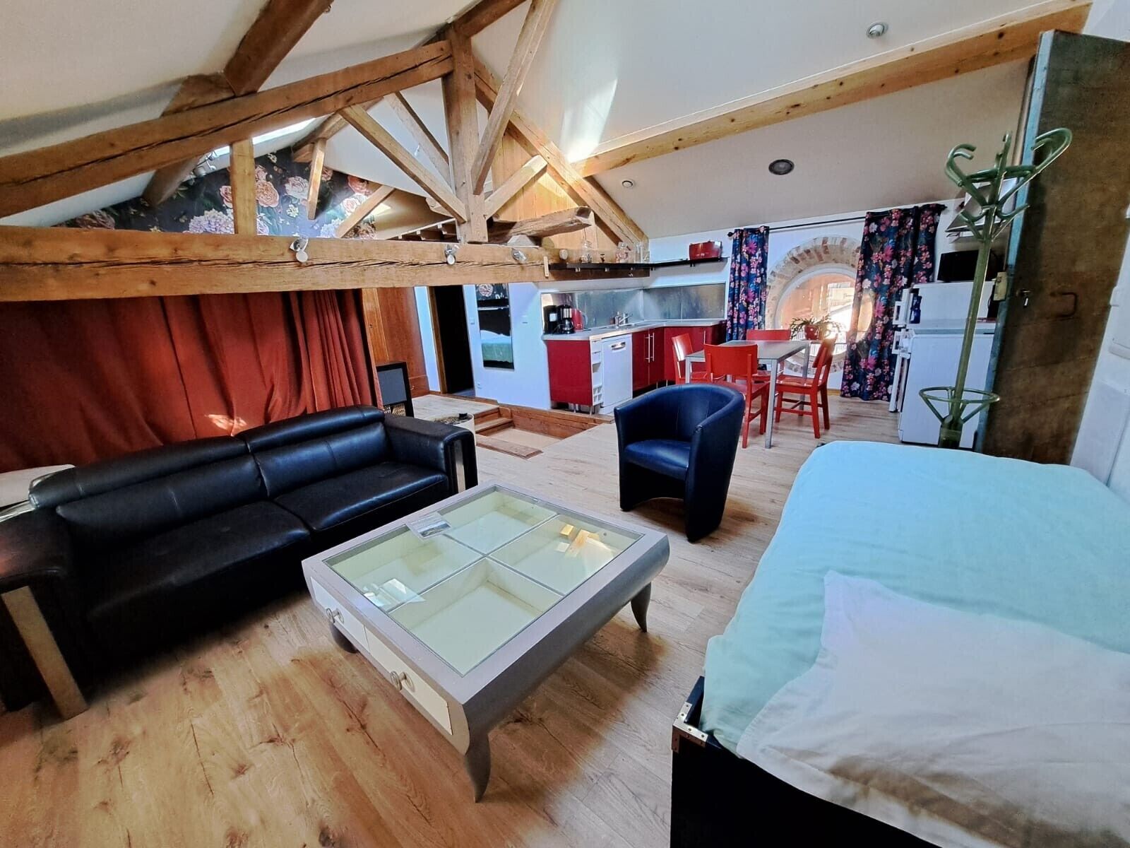 A loft-style living area with a wooden ceiling, a black leather sofa, a glass coffee table, a blue armchair, and a bed with a white cover.