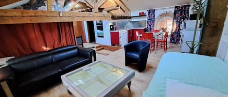 Un espacio tipo loft con techo de madera, un sofá de cuero negro, una mesa de centro de vidrio, un sillón azul y una cama con cubierta blanca.