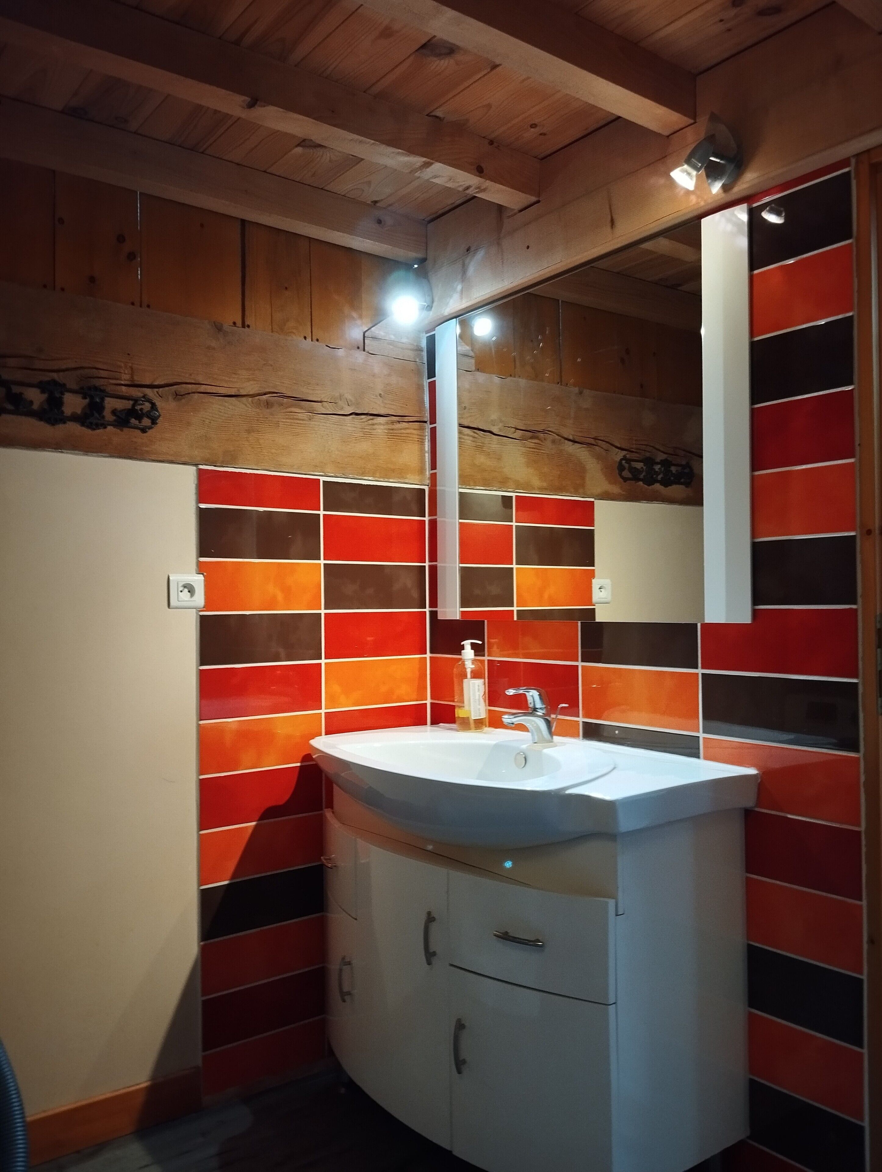 A bathroom with a wooden ceiling, a white sink with a mirror above it, and multicolored tiles on the walls.