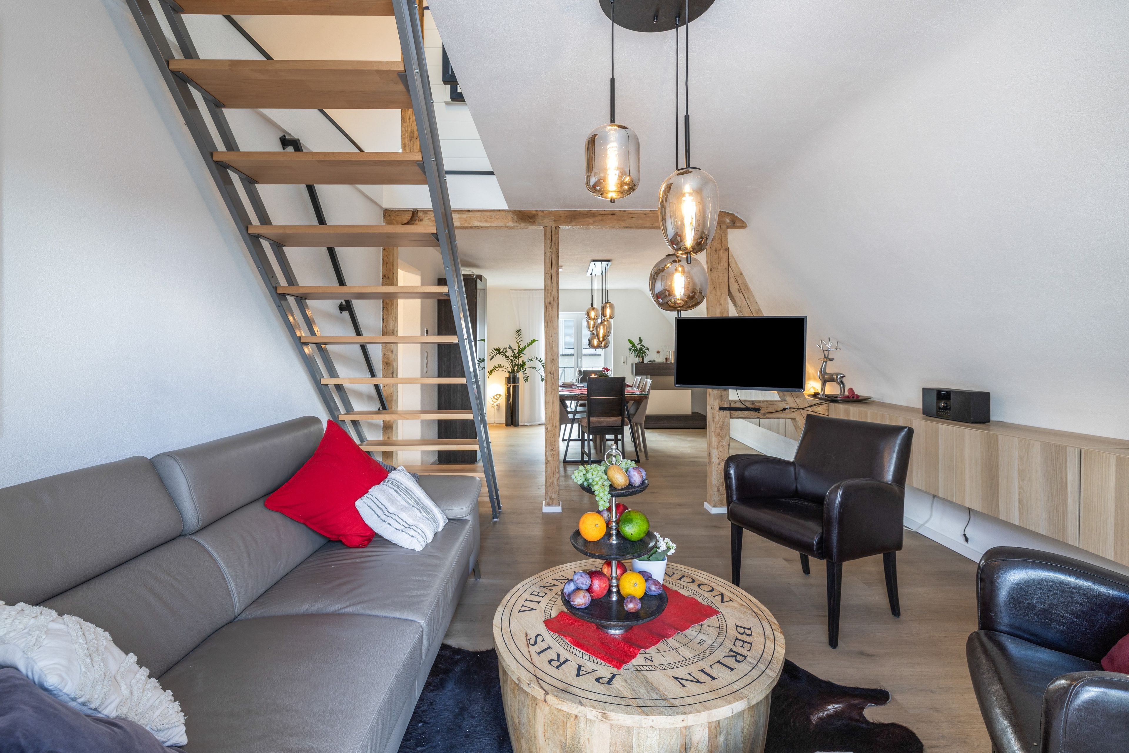 Ein modernes Wohnzimmer mit einem grauen Sofa, einem Couchtisch mit Obst, einer Treppe und einem Flachbildfernseher.