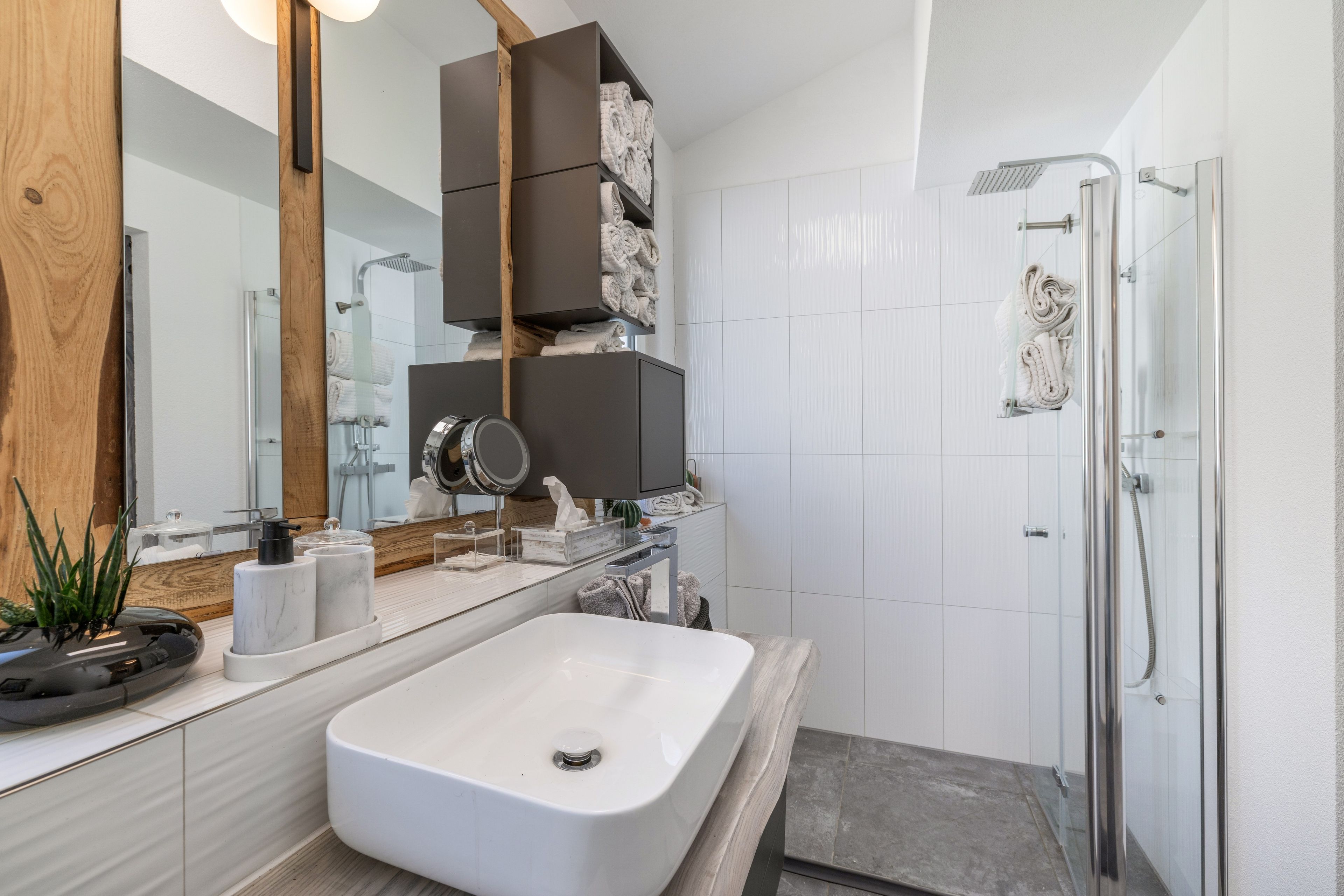 Ein modernes Badezimmer mit einem großen Spiegel, einem weißen Waschbecken, einer Dusche und einem Holzkästen über dem Spiegel.