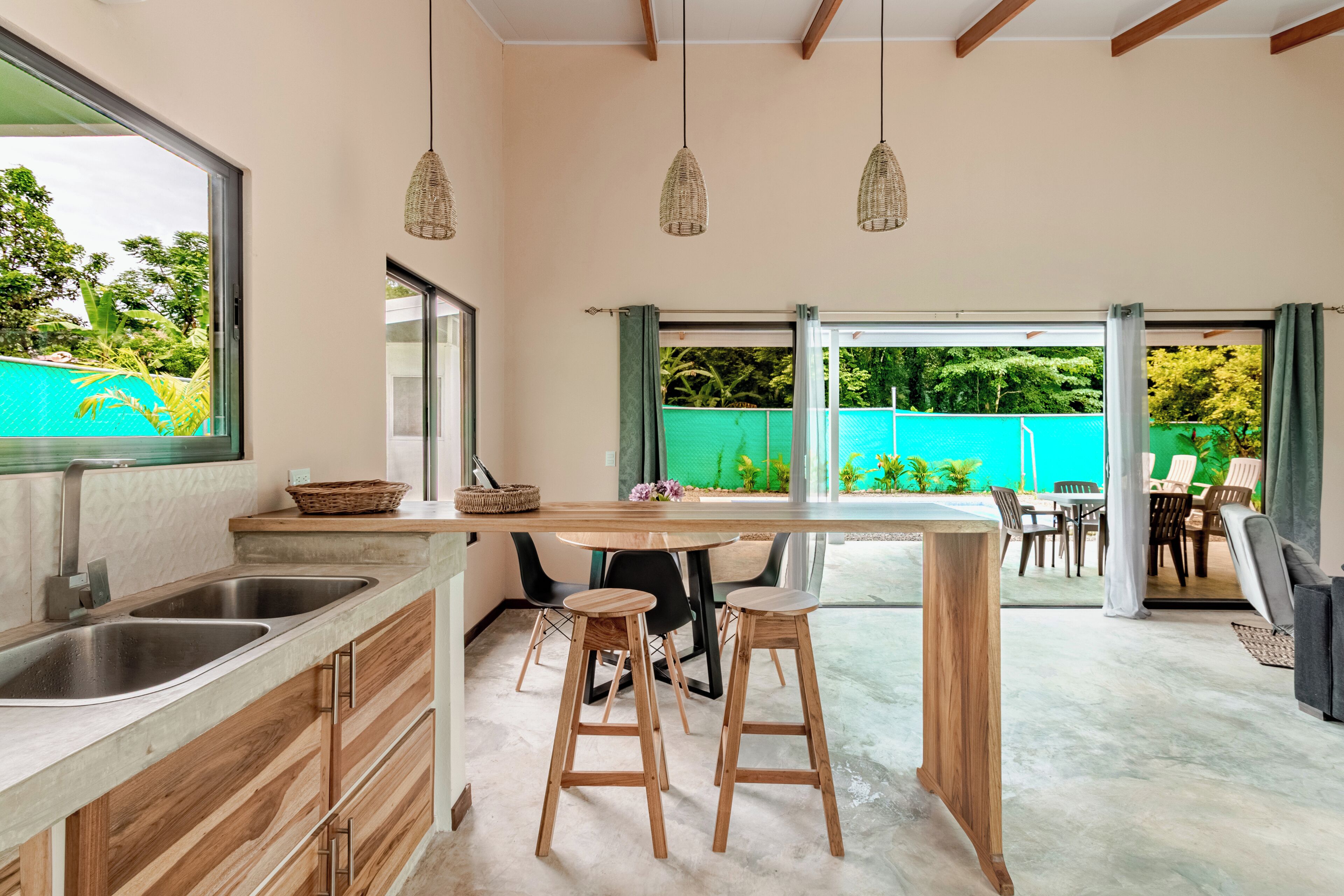A modern kitchen with a large island, bar stools, and a dining area with a view of a pool and greenery.