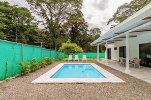 Ein rechteckiger Pool mit weißer Liegefläche, umgeben von einem grünen Zaun und Kies. Vier weiße Stühle und ein Tisch befinden sich unter einem Vordach.