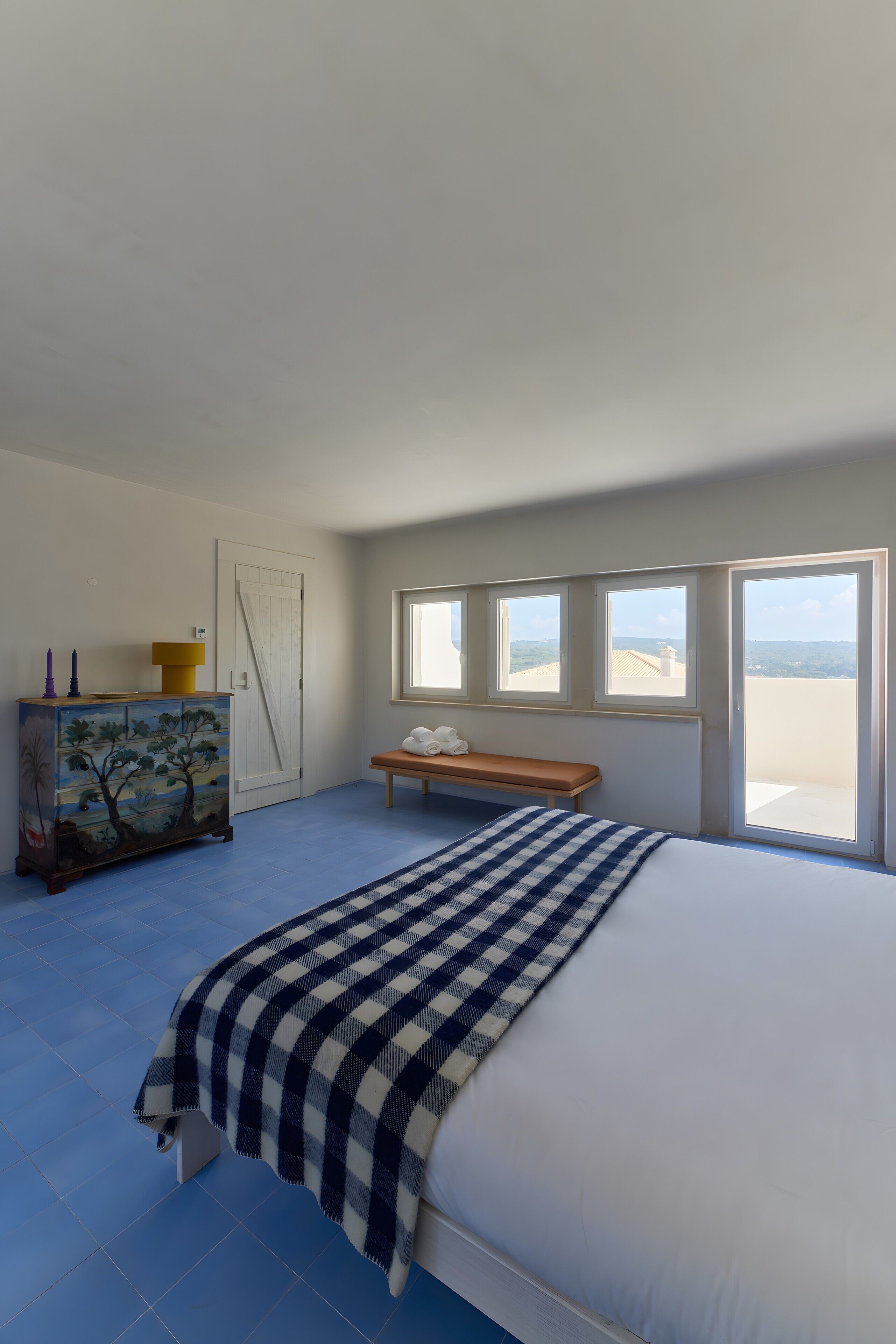 A bedroom with a large bed, a bench, a dresser, and a view of the landscape through the windows.