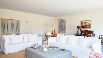 Ein Wohnzimmer mit zwei weißen Sofas, einem Couchtisch und einer Lampe. An den Wänden befinden sich Bilder und ein kleiner Tisch mit Dekorationsobjekten.