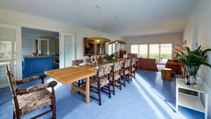 Un comedor espacioso con una larga mesa de madera y sillas tapizadas, una cocina con azulejos azules y un amplio ventanal que proporciona luz natural.