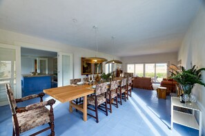 A spacious dining area with a long wooden table and upholstered chairs, a kitchen with blue tiles, and a large window providing natural light. - Pool Villa Stipa - Praia Grande (Colares)