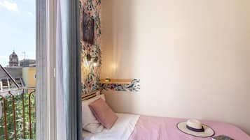 Un dormitorio con una cama, una ventana, vistas de un edificio y un sombrero sobre la cama.