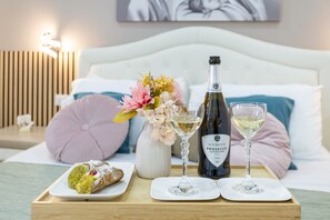 A bed with a white headboard, a wooden nightstand with a lamp, a tray with a bottle of wine, two glasses, a dessert, and a vase with flowers.