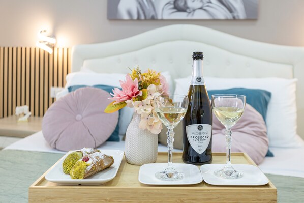 A bed with a white headboard, a wooden nightstand with a lamp, a tray with a bottle of wine, two glasses, a dessert, and a vase with flowers.