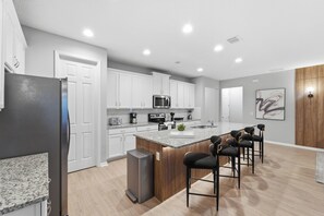 A modern kitchen with white cabinets, a granite countertop, and a central island with bar stools. - 7BR Themed Home | Solara Resort + Game, Cinema (Kissimmee)