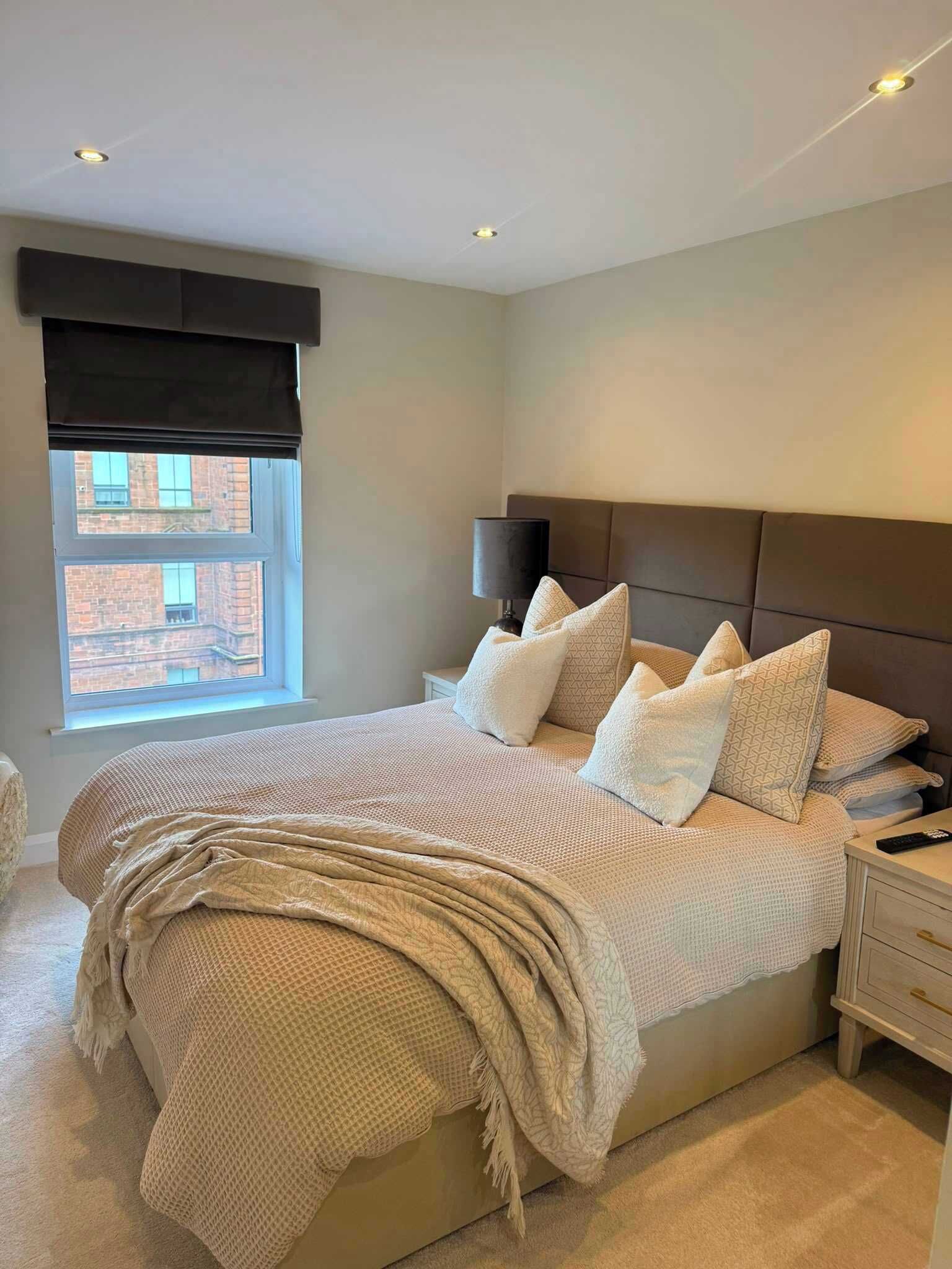 A bedroom with a large bed, a nightstand, a lamp, and a window with a view of a building.