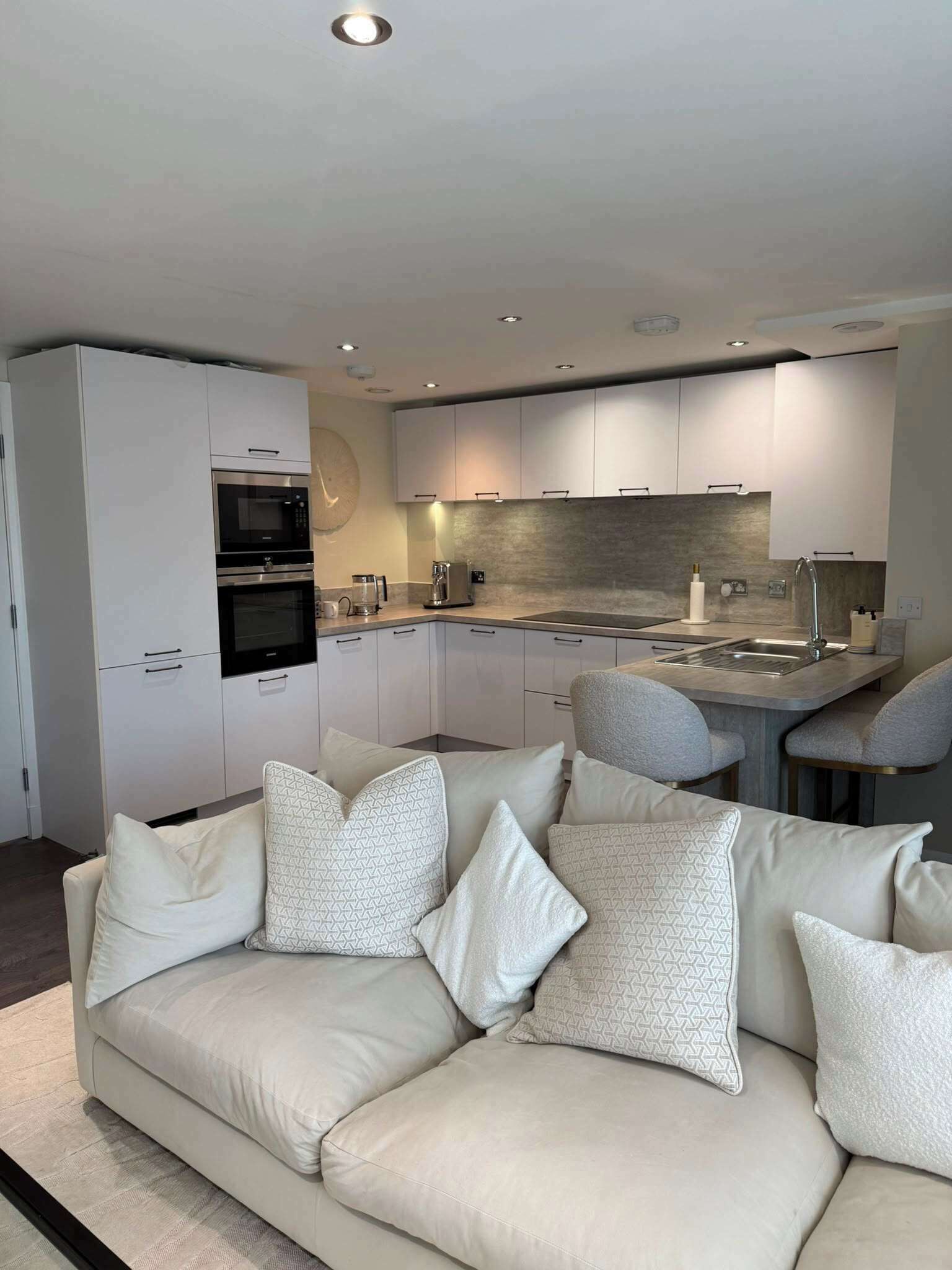 A modern kitchen with built-in appliances, a dining area with a grey sofa, and a large window.
