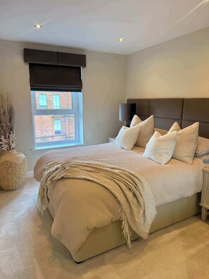 Une chambre à coucher moderne avec un grand lit, une fenêtre donnant sur un immeuble en briques et une tête de lit de couleur sombre.