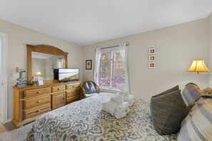 Une chambre à coucher avec un lit, une commode, un téléviseur, une fenêtre avec des rideaux et une lampe.