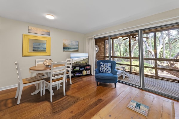 A room with a wooden floor, a dining table with chairs, a blue armchair, a television, and a large window with a view of trees. - Kiawah Exclusives | 4276 Mariners Watch Villa (Kiawah Island)