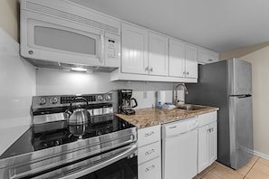 A compact kitchen with white cabinets, a stainless steel stove, a microwave, a refrigerator, and a granite countertop. - Kiawah Exclusives | 4276 Mariners Watch Villa (Kiawah Island)