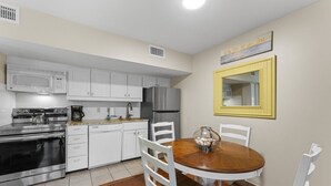 Cocina compacta con armarios blancos, un refrigerador de acero inoxidable, microondas y estufa. Hay una mesa de comedor con sillas blancas y un cuenco decorativo.