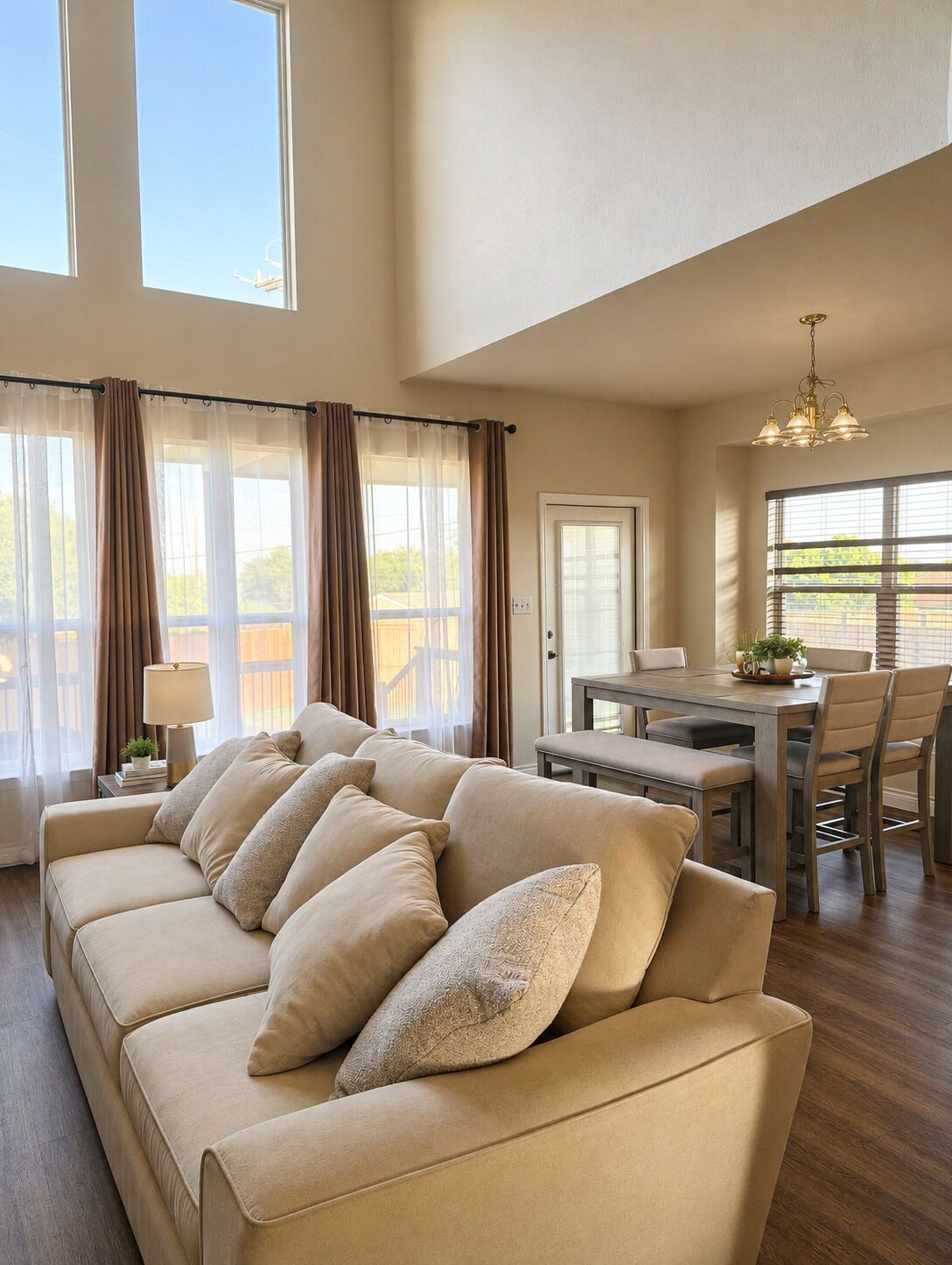 A spacious living room with a beige sofa, a dining table with chairs, and large windows with curtains.