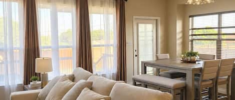 A spacious living room with a beige sofa, a dining table with chairs, and large windows with curtains.