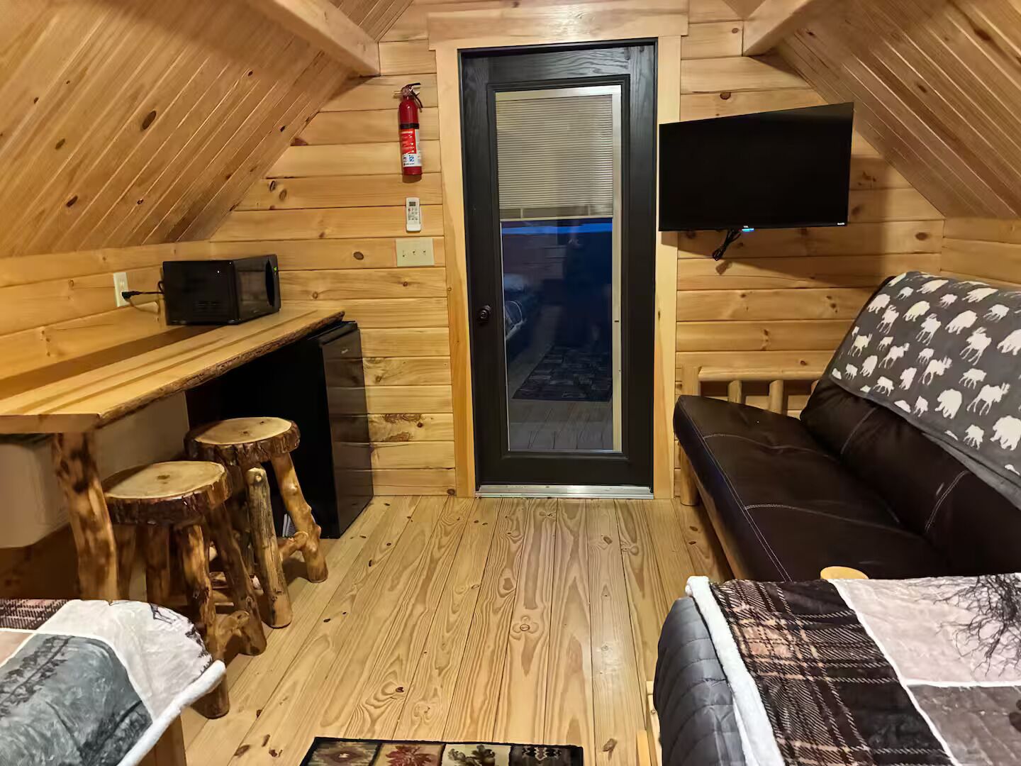 Habitación en ático con techo inclinado de madera, un sofá, una pequeña mesa con taburetes, un televisor montado en la pared y una puerta que ofrece vistas al exterior.