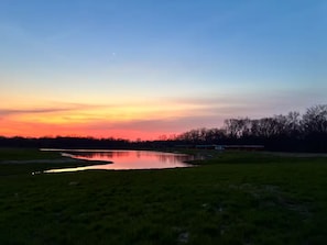 A serene landscape with a body of water reflecting the sunset sky, surrounded by grassy fields and a line of trees.