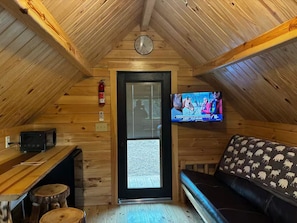 A cozy attic room with a wooden ceiling, a television mounted on the wall, a black leather sofa, and a wooden bench.