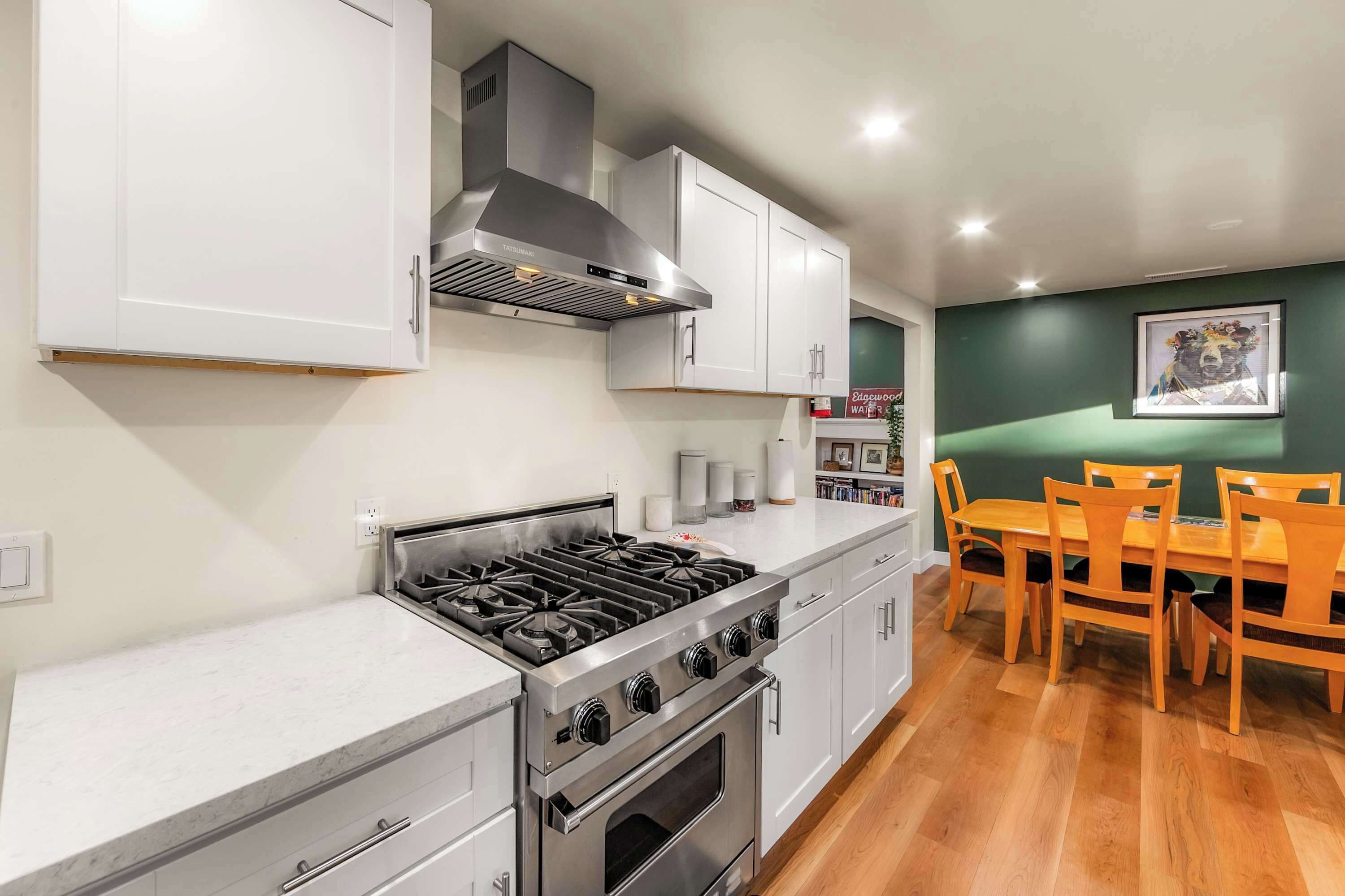 Cocina moderna con estufa a gas, horno y armarios blancos. Hay una mesa de comedor con sillas y una pared verde con un cuadro enmarcado.