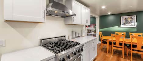 Cocina moderna con estufa a gas, horno y armarios blancos. Hay una mesa de comedor con sillas y una pared verde con un cuadro enmarcado.