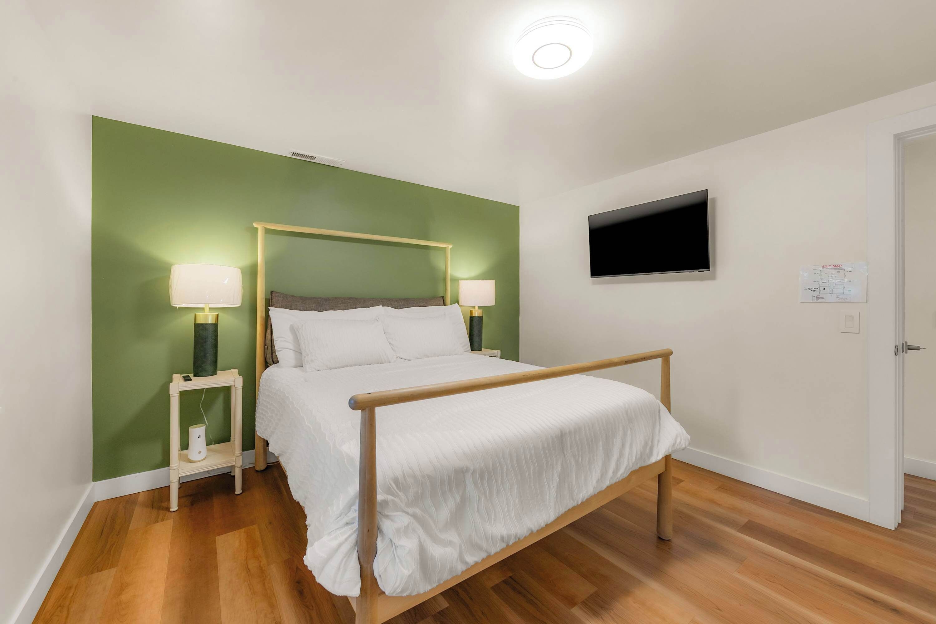 Uma cama de solteiro com estrutura de madeira e roupa de cama branca, ladeada por duas mesas de cabeceira com luminárias, em um quarto com paredes verdes e piso de madeira.