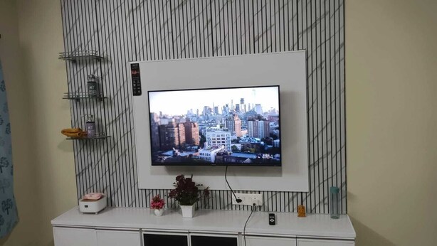 Televisão fixada na parede com paisagem urbana na tela, um console branco abaixo e uma planta sobre o console.