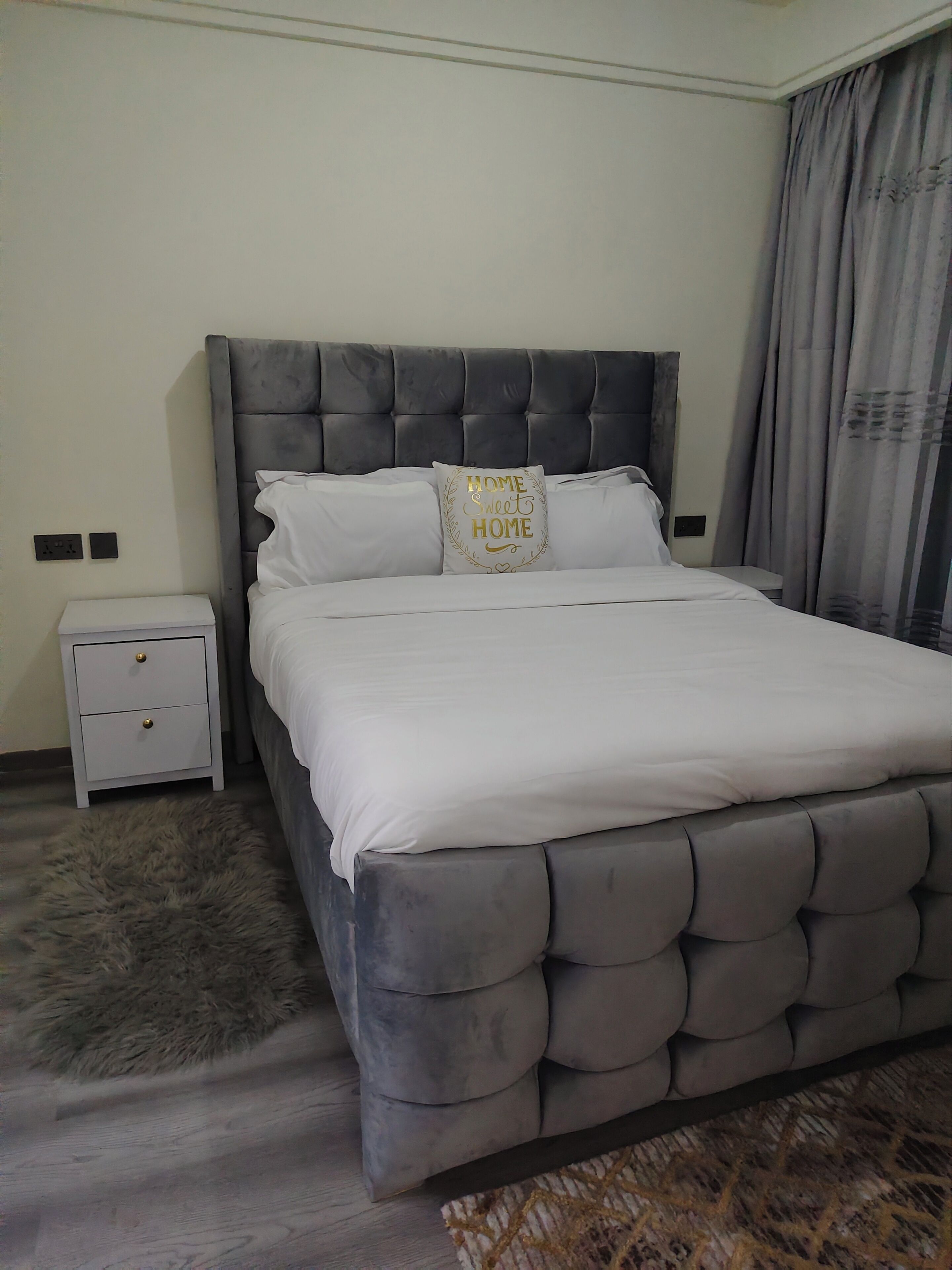 A bedroom with a tufted headboard, a white bedspread, a fluffy rug, a nightstand, and a grey curtain.