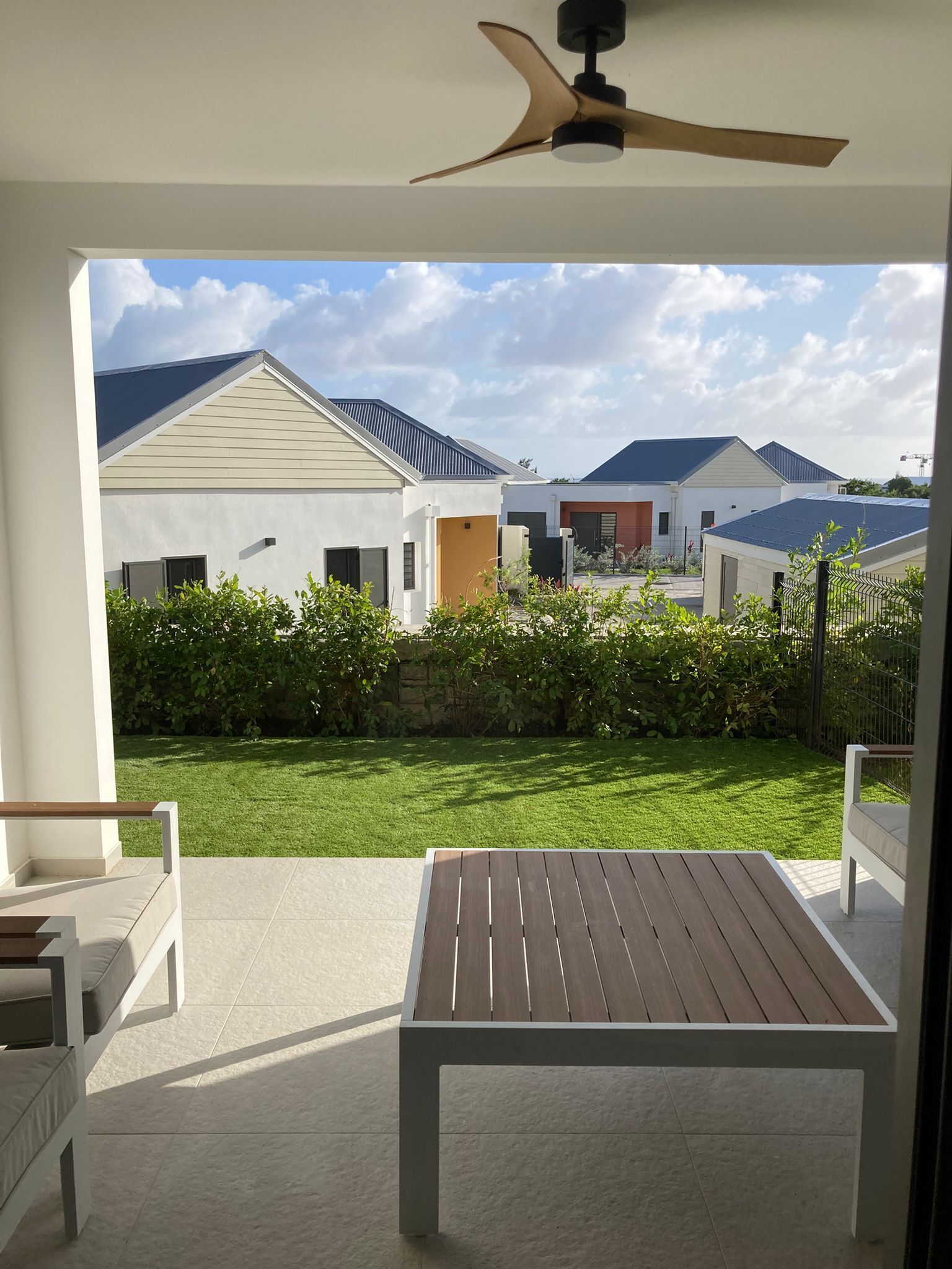Eine moderne Terrasse mit Holztisch und -stühlen, einem Deckenventilator und Blick auf eine Wohngegend mit Häusern und einem gepflegten Rasen.