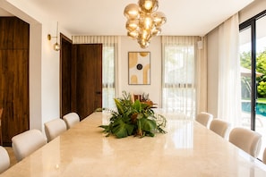 A modern conference room with a marble table, white chairs, a potted plant centerpiece, a wall-mounted abstract painting, and a large window with a view of a pool. - El Bosque II Casa de Campo (La Romana)