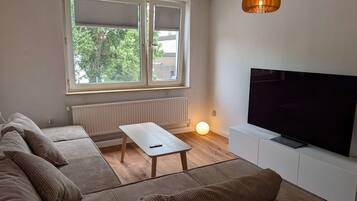Ein Wohnzimmer mit einer Sofagarnitur, einem Couchtisch, einem Fernseher auf einem Lowboard und einem Fenster mit Jalousien.