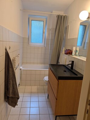 A bathroom with a bathtub, a toilet, a sink with a mirror, and a towel rack. - Stylish 1-Bedroom Apartment with Balcony | Close to the City Centre (Hannover)