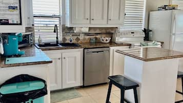 A kitchen with white cabinets, a dishwasher, a sink, and a countertop with a trash bin and a bar stool.