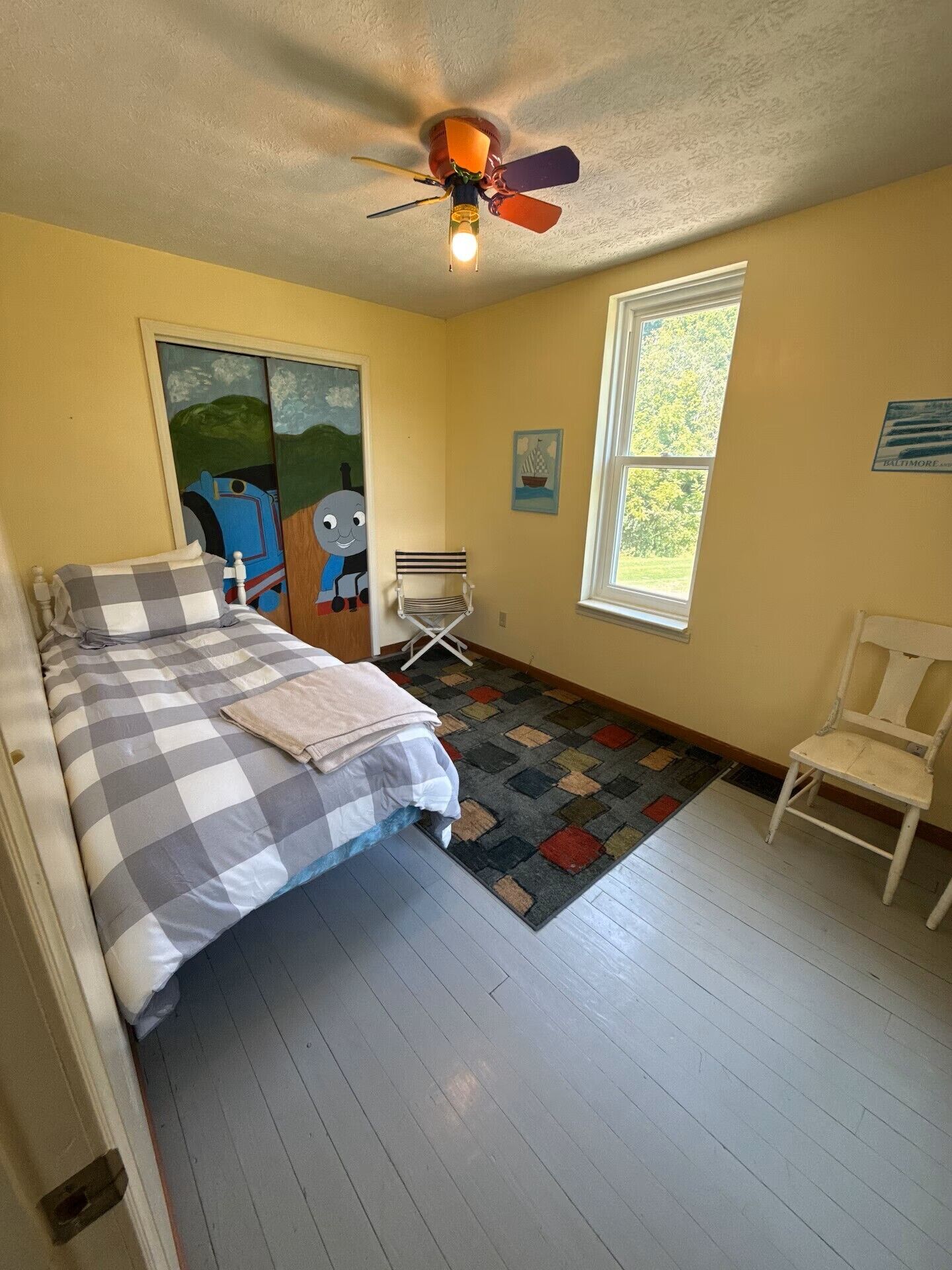 Un dormitorio con cama, una silla, un ventanal y un ventilador de techo.