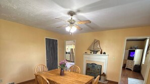 Uma sala de jantar com mesa e cadeiras de madeira, um ventilador de teto e lareira.