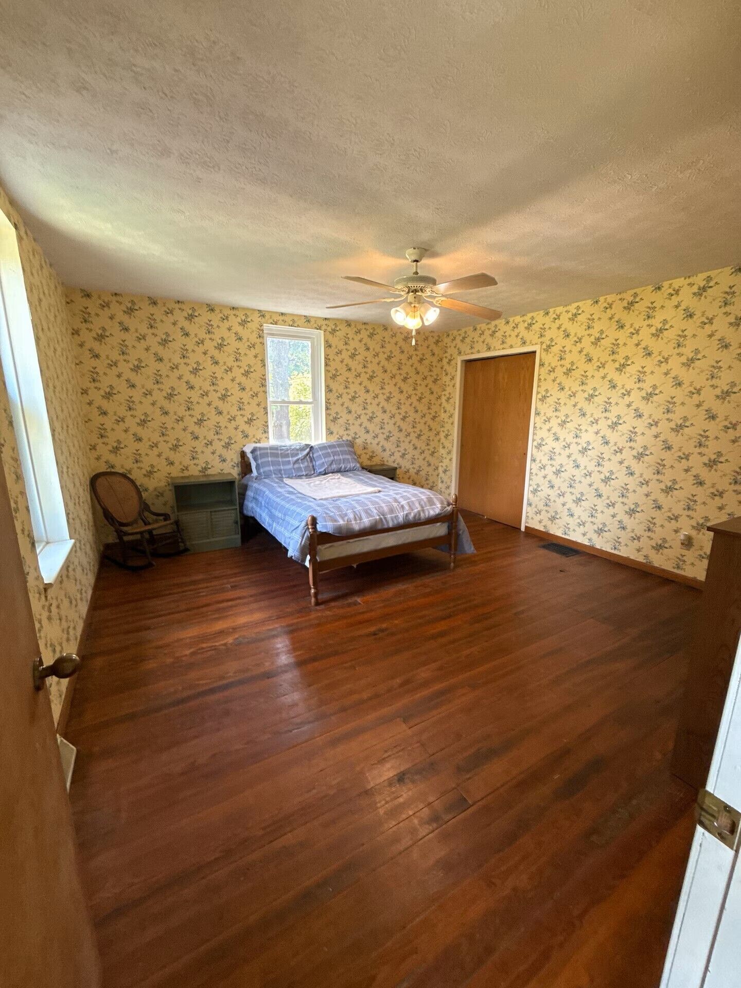A bedroom with a wooden floor, a bed with a blue and white checkered bedspread, a ceiling fan, and floral wallpaper.