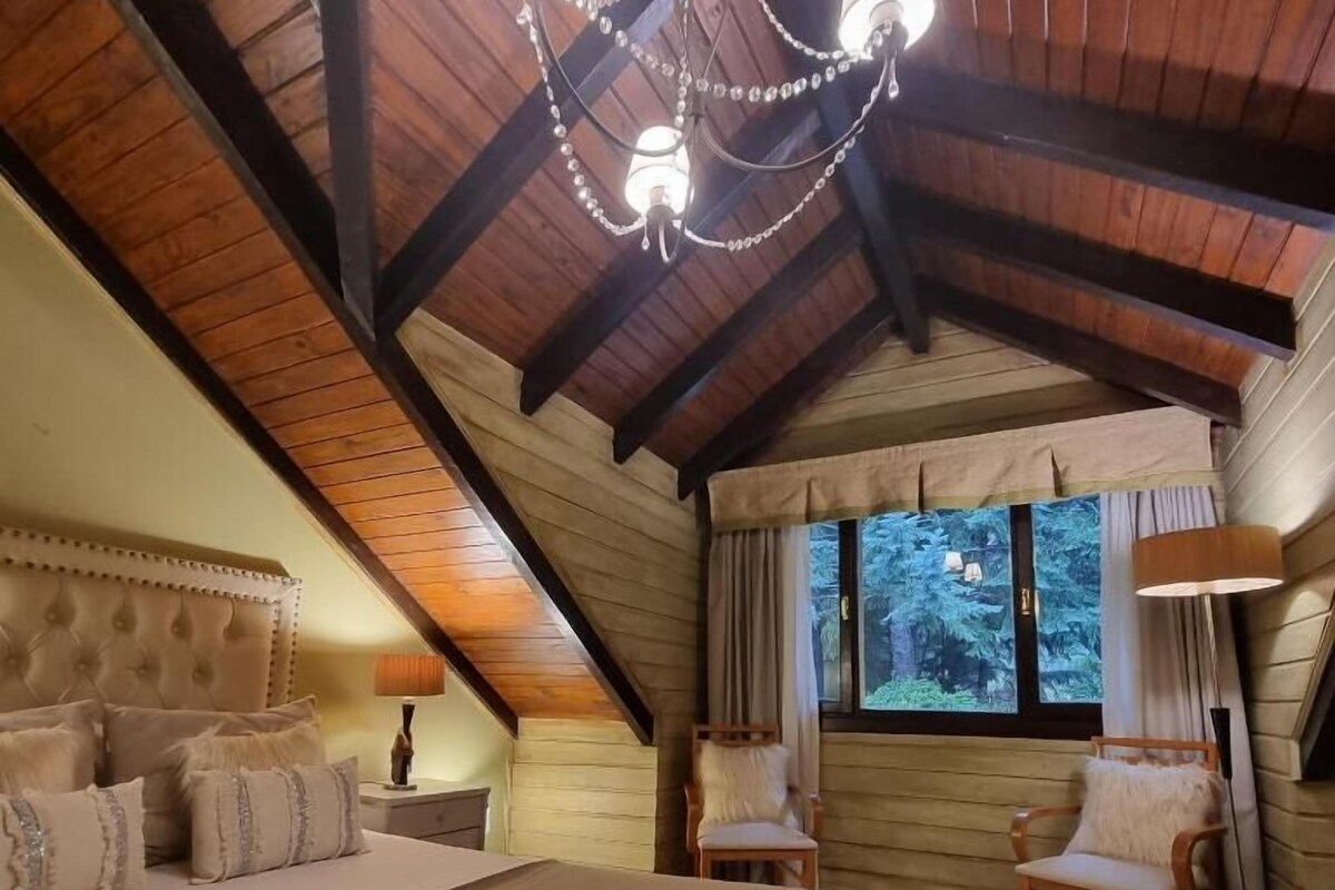 A bedroom with a large bed, wooden beams, a chandelier, and a window with a view of trees.