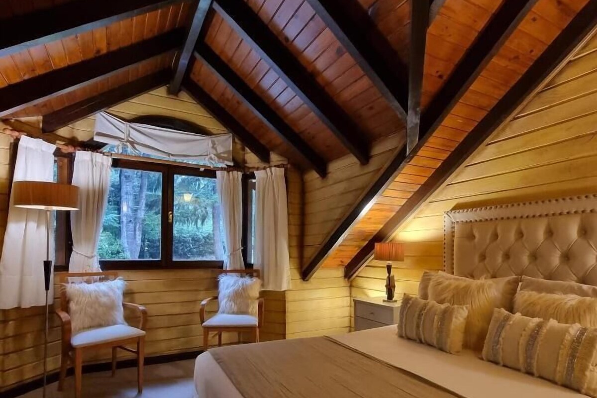 A bedroom with a large bed, two chairs, a window with curtains, and a wooden ceiling.