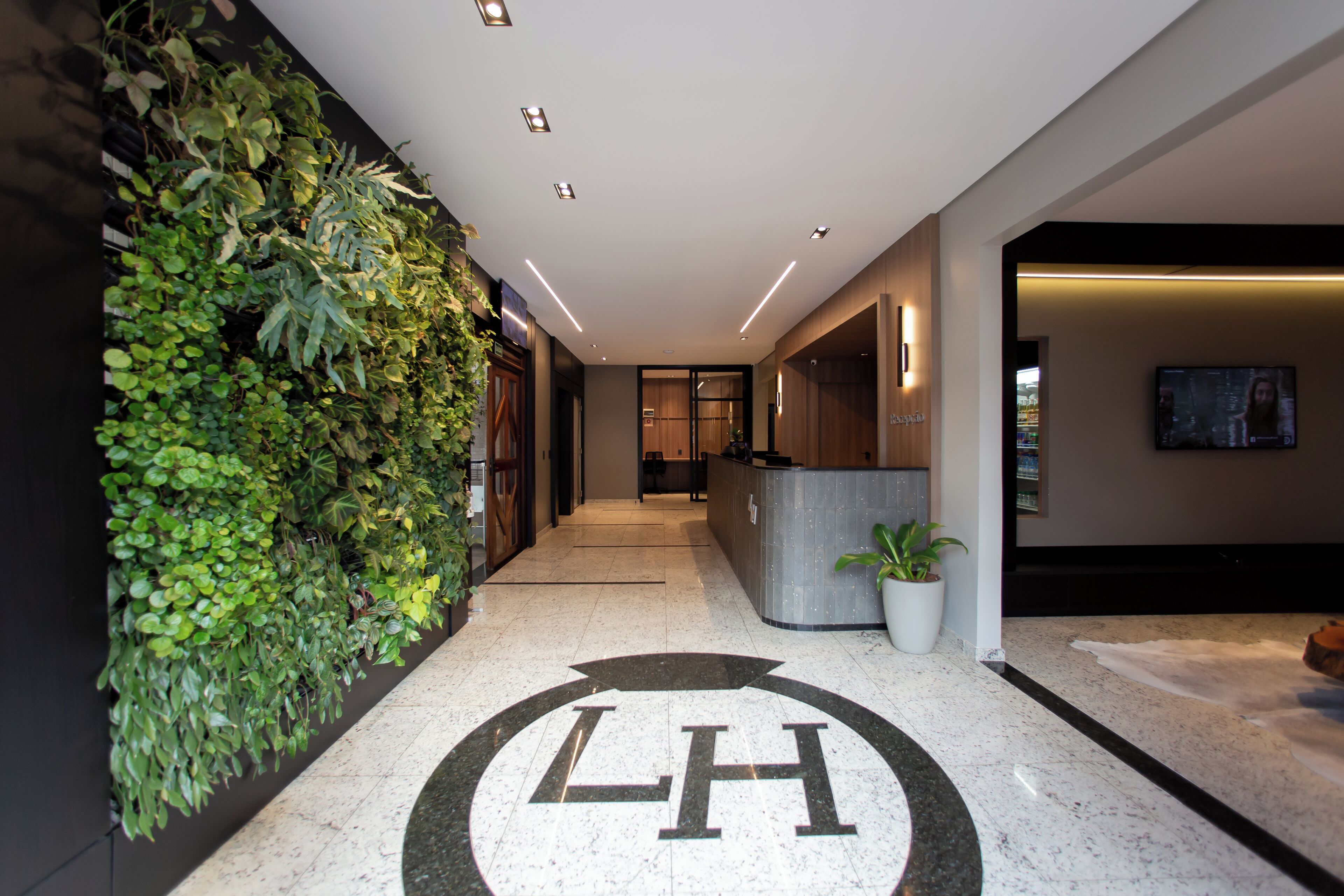 A hotel reception area with a reception desk, a large logo on the floor, a wall with greenery, and a television screen.