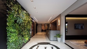 Zona de recepción de hotel con mostrador, un gran logotipo en el suelo, una pared con plantas y una pantalla de televisión.
