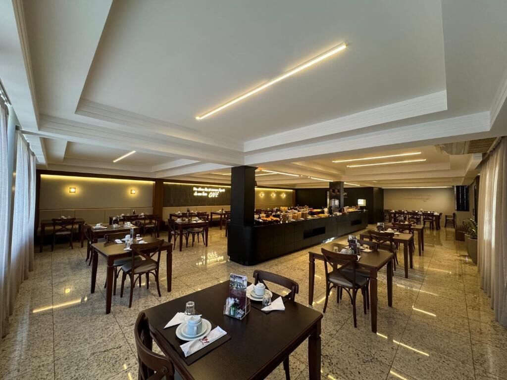 A spacious hotel dining area with multiple tables and chairs, a buffet station, and a modern ceiling design.