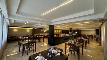 A spacious hotel dining area with multiple tables and chairs, a buffet station, and a modern ceiling design.